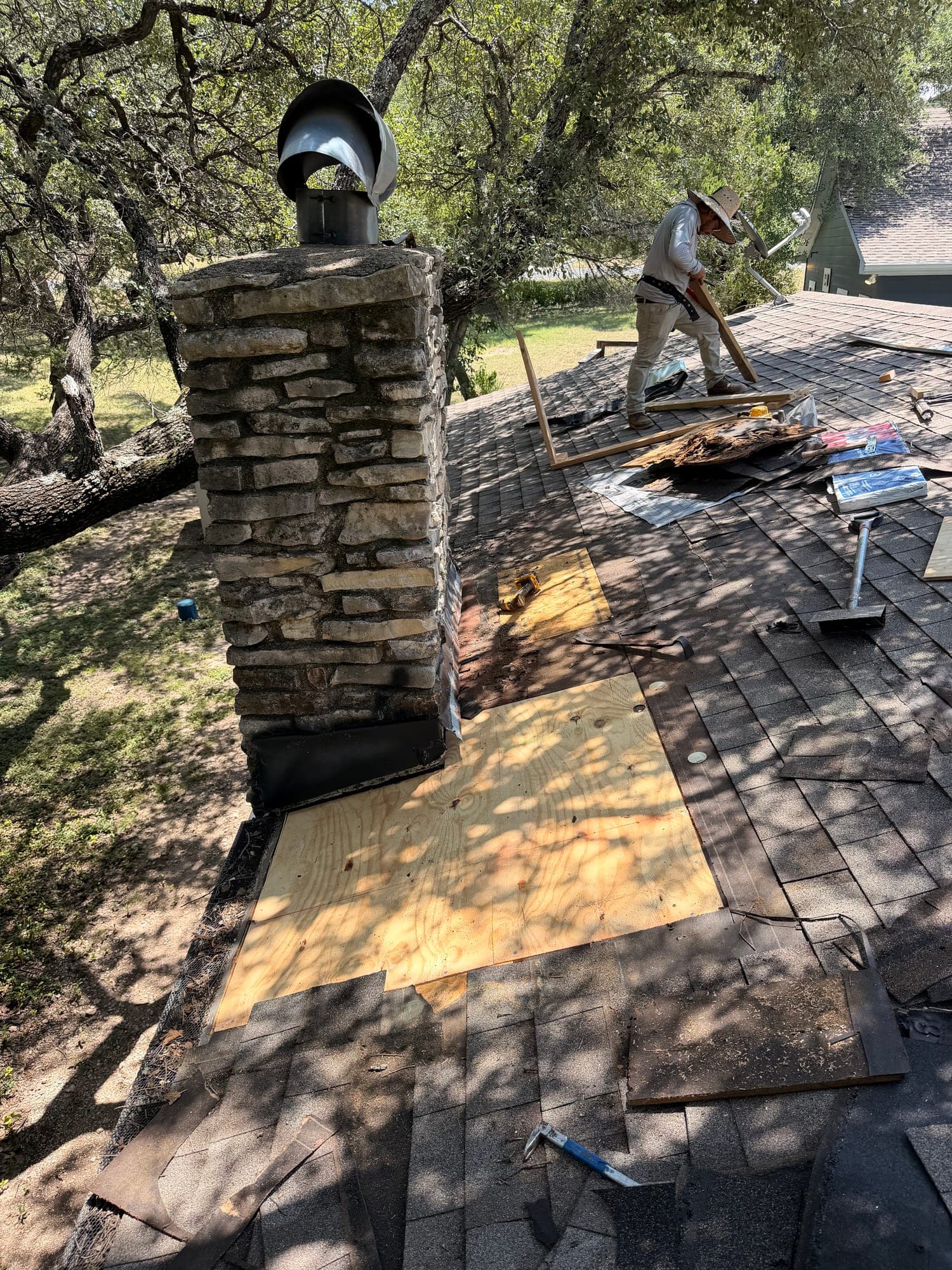 New plywood decking installed at the base of a stone chimney while tear-off continues up the slope behind it