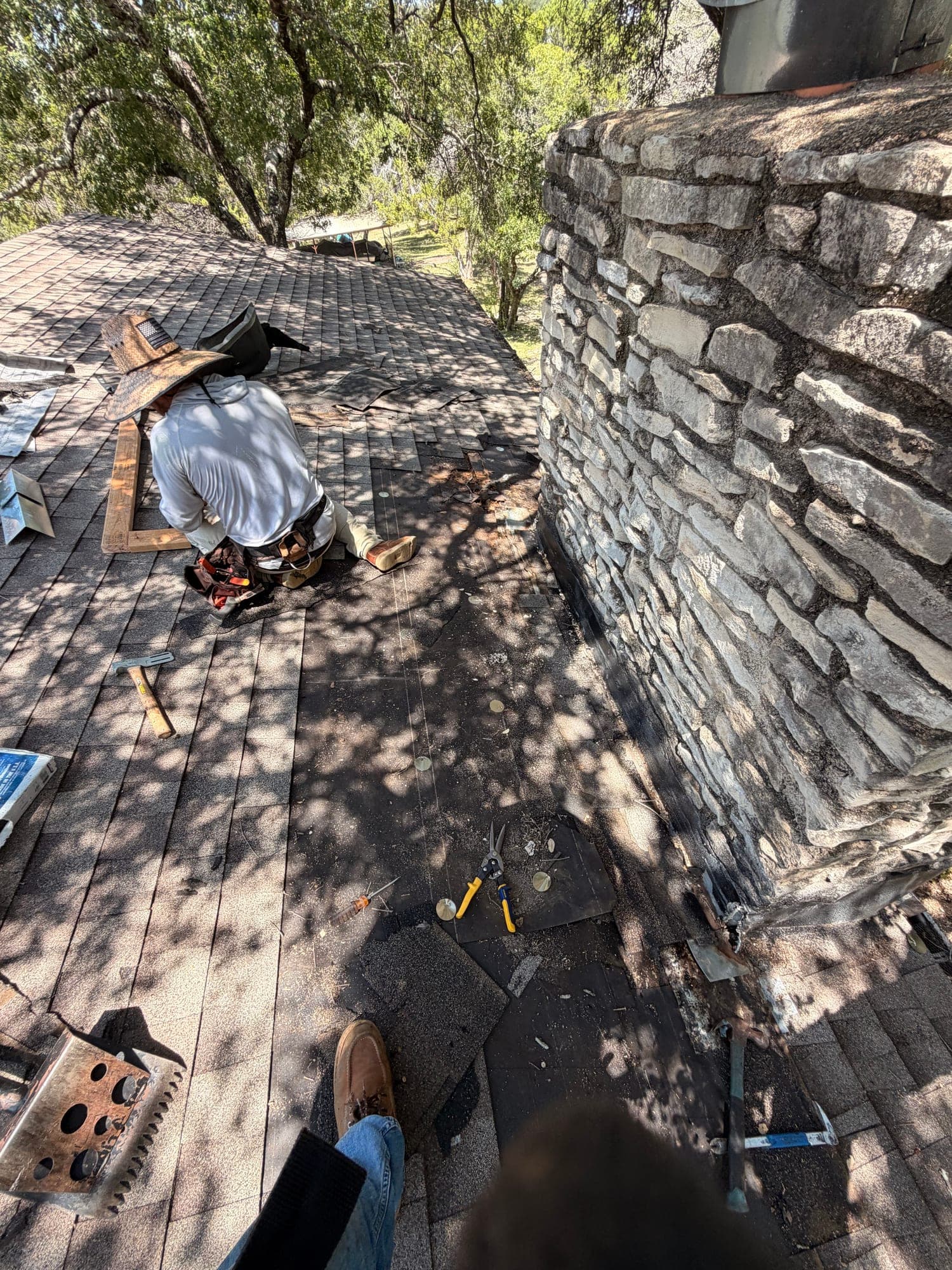 Roofer working beside a stone chimney during tear-off, shingles and felt stripped back to the deck around the base